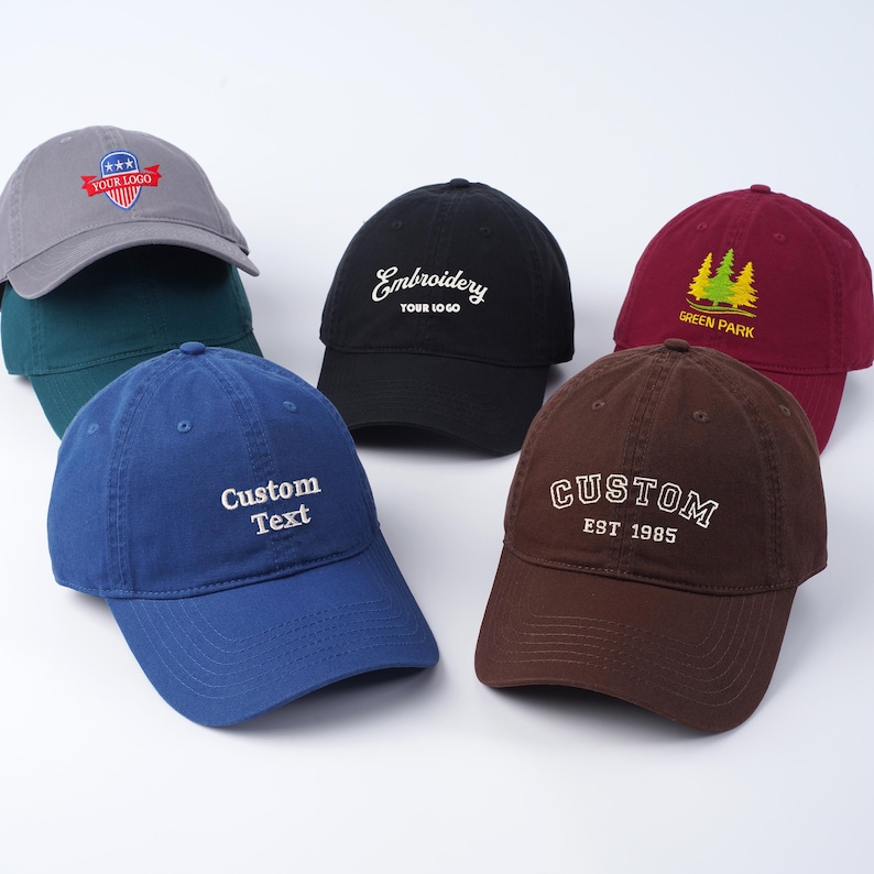 May include: Six different baseball caps with various embroidered designs. The caps are in different colors, including gray, black, maroon, blue, and brown. The designs include a star with the words "Your Logo", "Embroidery Your Logo", "Green Park" with a tree design, "Custom Text", and "CUSTOM EST 1985".