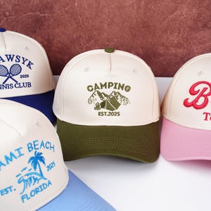 May include: A collection of baseball caps with various designs. One cap features "CAMPING" with a mountain graphic and "EST.2025". Other caps display "AWSYK TENNIS CLUB 2025", "MIAMI BEACH FLORIDA 2025", and a script "B". The caps have different color combinations.