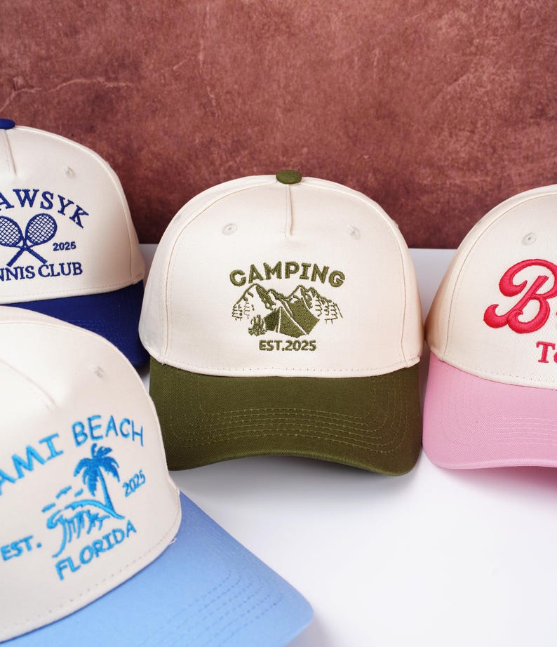May include: A collection of baseball caps with various designs. One cap features "CAMPING" with a mountain graphic and "EST.2025". Other caps display "AWSYK TENNIS CLUB 2025", "MIAMI BEACH FLORIDA 2025", and a script "B". The caps have different color combinations.