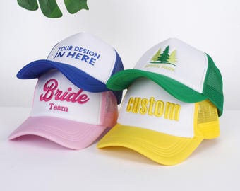 Puede incluir: Tres gorras de camionero con diferentes diseños. La primera gorra es azul y blanca con el texto "YOUR DESIGN IN HERE". La segunda gorra es rosa y blanca con el texto "Bride Team". La tercera gorra es amarilla, verde y blanca con el texto "Custom" y un diseño de árbol verde.