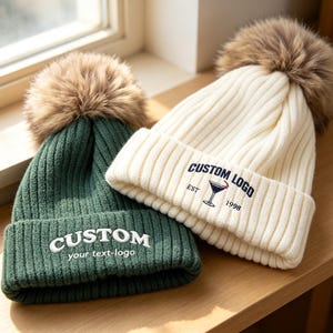 Puede incluir: Un gorro de punto acanalado verde oscuro con un pompón de piel sintética marrón y texto bordado en blanco CUSTOM your text-logo, junto a un gorro de punto acanalado crema con pompón marrón y texto azul marino CUSTOM LOGO EST 1998.