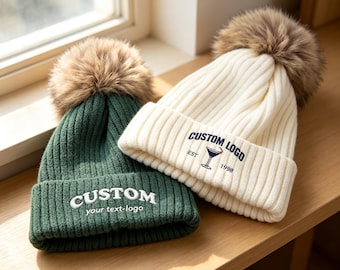 May include: A dark green ribbed knit beanie with a brown faux fur pom-pom and white embroidered text reading CUSTOM your text-logo, alongside a cream ribbed knit beanie with a brown faux fur pom-pom and navy text reading CUSTOM LOGO EST 1998.