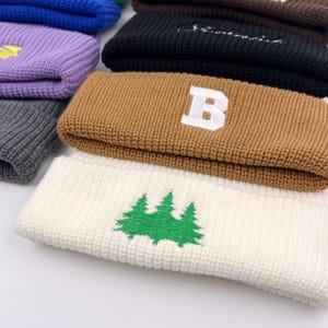 May include: A stack of knitted beanies in various colors, including brown, white, black, purple, and gray. The brown beanie has a white letter "B" embroidered on it. The white beanie has a green embroidered design of three pine trees.