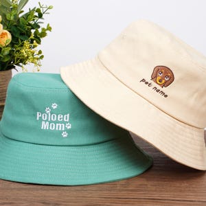 Pu&ograve; includere: Due cappelli a secchio. Uno &egrave; color crema con una grafica di bassotto marrone e la scritta "pet name". L'altro &egrave; verde acqua con la scritta "Poloed Mom" e impronte di zampe. Entrambi i cappelli hanno un'ampia tesa.