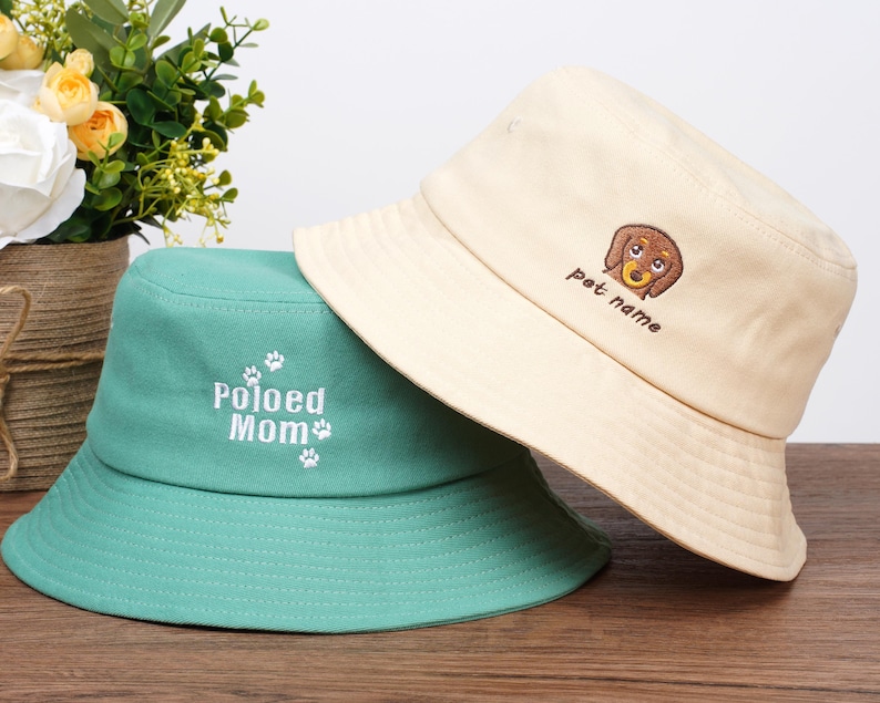 Pu&ograve; includere: Due cappelli a secchio. Uno &egrave; color crema con una grafica di bassotto marrone e la scritta "pet name". L'altro &egrave; verde acqua con la scritta "Poloed Mom" e impronte di zampe. Entrambi i cappelli hanno un'ampia tesa.