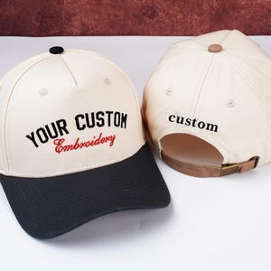 May include: Two baseball caps with custom embroidery. The caps have a cream-colored crown and a black brim. The text "YOUR CUSTOM Embroidery" is embroidered on the front in black and red. The back of one cap has the word "custom" embroidered in black.