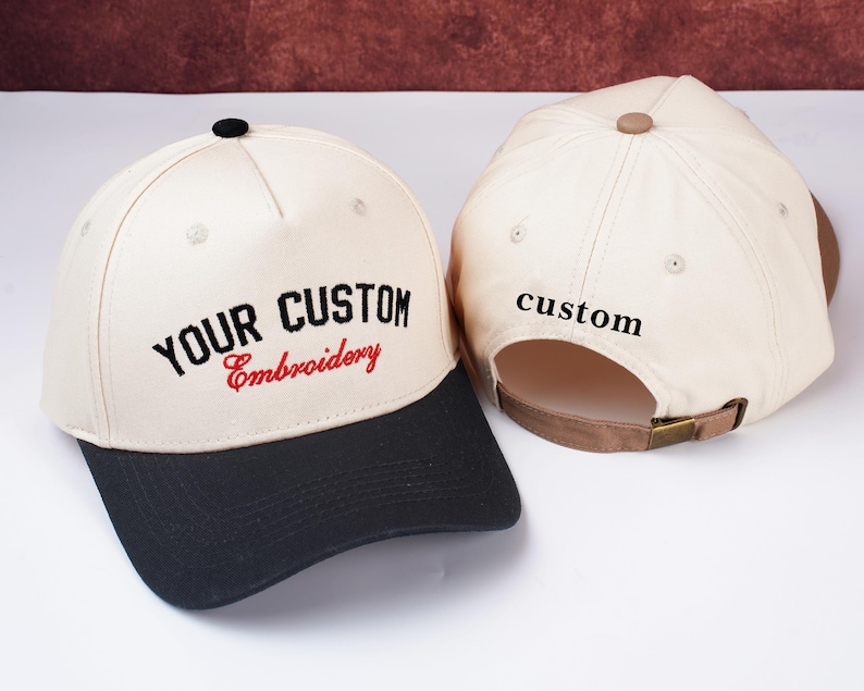 May include: Two baseball caps with custom embroidery. The caps have a cream-colored crown and a black brim. The text "YOUR CUSTOM Embroidery" is embroidered on the front in black and red. The back of one cap has the word "custom" embroidered in black.