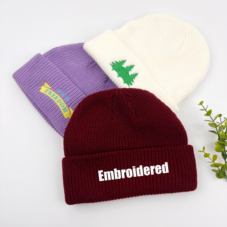 May include: Three knitted beanies in different colors. One is purple with the word "FREEDOM" embroidered on it, another is white with a green pine tree design, and the third is burgundy with the word "Embroidered" in white.