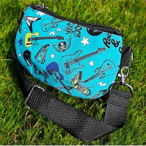 May include: A turquoise crossbody bag with a black strap, featuring a rock and roll theme. The bag is patterned with electric guitars, stars, and the words "Rock Star". The bag has a black zipper and silver hardware.