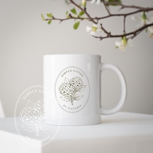 Op de afbeelding: Witte keramische mok met een bloemenontwerp en de tekst "SIMPLE LIVING by VICTORIA". De mok heeft een handvat en een cirkelvormig logo met een botanische illustratie van bloemen. Een takje bloemen staat op de achtergrond.