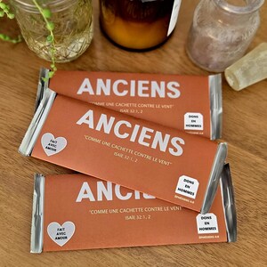 Puede incluir: Tres barras de color naranja con la palabra "ANCIENS" impresa en blanco. Las barras tienen texto en francés y están empaquetadas en papel de aluminio plateado. Otros artículos incluyen frascos de vidrio y una vela.