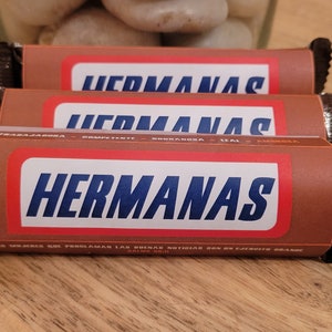 Peut inclure: Trois barres de chocolat brun avec le mot "HERMANAS" imprimé en bleu et blanc sur le devant. Les barres sont enveloppées dans du papier d'aluminium brun avec un bord dentelé.