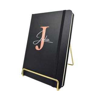 Könnte beinhalten: Ein schwarzes, ledergebundenes Notizbuch mit einem goldenen Metallständer. Das Notizbuch hat einen großen pinkfarbenen Buchstaben "J" und den Namen "Julia" in weißer Kursivschrift.