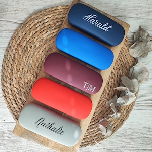 Könnte beinhalten: Fünf personalisierte Brillenetuis auf einem Holzständer. Die Etuis sind in verschiedenen Farben: grau, rot, kastanienbraun, blau und dunkelblau. Die Namen "Nathalie", "T/M" und "Harald" sind auf die Etuis gedruckt.
