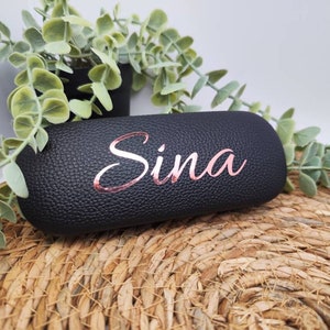 Könnte beinhalten: Schwarzes Brillenetui aus Kunstleder mit dem Namen "Sina" in roségoldenen Lettern geprägt.