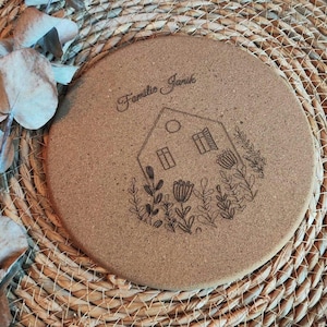 Könnte beinhalten: Ein runder Korkuntersetzer mit einem Haus- und Blumendesign. Der Text "Familia Janik" ist in den Untersetzer graviert.