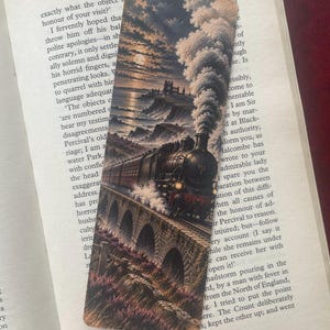 May include: A rectangular bookmark featuring a detailed illustration of a steam locomotive traveling across a stone viaduct at sunset. The train emits a thick plume of smoke against a cloudy sky, with a distant castle visible on the rocky coastline.