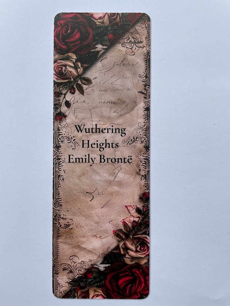 May include: A rectangular bookmark featuring a vintage parchment background with cursive handwriting, ornate dark borders, and clusters of deep red and cream-colored roses at the top and bottom corners. The text reads Wuthering Heights Emily Bront&euml;.