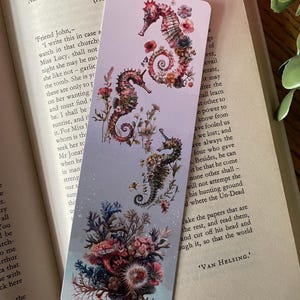 May include: A rectangular bookmark with a light purple to blue gradient background featuring three seahorses adorned with colorful floral elements and a dense cluster of coral and sea anemones at the bottom edge of the bookmark.