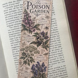 May include: A rectangular paper bookmark with a vintage beige background featuring botanical illustrations of poisonous plants. The text reads The Poison Garden, Foxglove, Nightshade, Hemlock, and Wolfsbane with their scientific names.