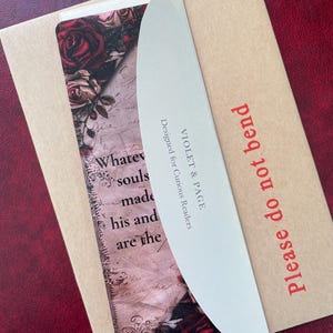 May include: A bookmark featuring a floral rose pattern and vintage script text, partially covered by a curved white card with the text VIOLET & PAGE Designed for Curious Readers, all resting on a tan envelope labeled Please do not bend.
