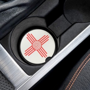 May include: A white car cup holder with a red and white circular coaster featuring the symbol of the state of New Mexico. The symbol is a red Zia sun symbol with four rays.