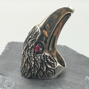 May include: A silver ring shaped like a raven's head with a red gemstone eye.