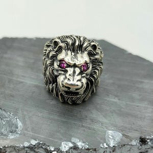 Può includere: Un anello in argento a forma di testa di leone con occhi di pietre preziose rosa. L'anello è dettagliato con pelliccia testurizzata e un'espressione feroce.