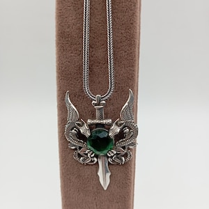 May include: A silver chain necklace with a pendant featuring a sword with a green gemstone surrounded by two dragons.