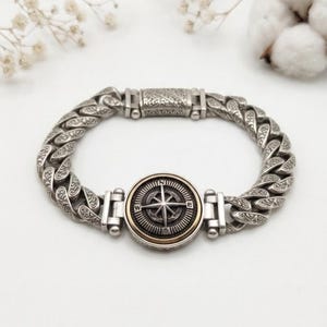 May include: Silver-coloured bracelet with a compass design. The bracelet features a thick chain link style with a circular compass centrepiece. The compass has a gold-coloured rim and a detailed design. The bracelet is on a white background.