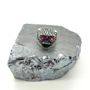 Op de afbeelding: Een zilveren schedelring met rode edelsteenogen. De ring heeft een textuurontwerp op het voorhoofd en de kaaklijn. De schedelring wordt gepresenteerd op een grijs, metalen oppervlak tegen een witte achtergrond.