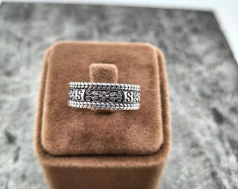 Puede incluir: Un anillo de plata con un diseño ornamentado con la letra "S" a cada lado. El anillo se muestra sobre una caja de terciopelo marrón, sobre un fondo neutro. Se destacan los intrincados detalles del anillo.
