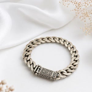 Op de afbeelding: Zilverkleurige armband met een brede schakelketting en een rechthoekige sluiting met een sierlijk ontwerp. De armband ligt op een witte ondergrond met een zachte, gedrapeerde stof. De schakels zijn ongeveer 1 cm breed.