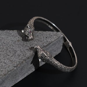 May include: A silver-toned cuff bracelet featuring detailed panther heads with red gemstone eyes. The bracelet is displayed on a gray stone surface against a dark background. The panther heads are intricately textured.