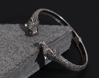 May include: A silver cuff bracelet with panther head terminals, each with a red gemstone eye. The bracelet is shown on a grey stone surface against a dark backdrop. The panther heads have detailed textures.