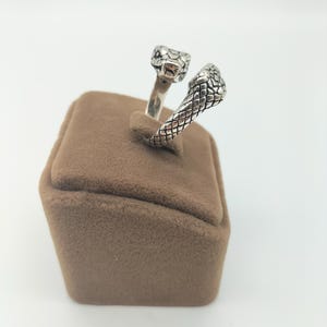 Op de afbeelding: Zilveren ring met een dubbel slangendesign. De slangenkoppen zijn naar voren gericht en de staarten zijn met elkaar verweven. De ring ligt op een bruine fluwelen displaystandaard.
