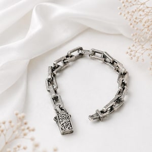 May include: A silver-colored chain bracelet with an ornate clasp, resting on a white satin surface. The bracelet features a series of rectangular links with detailed patterns. The clasp is rectangular with a decorative design.