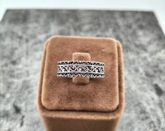 Puede incluir: Un anillo de plata con un intrincado diseño calado, con un patrón repetido de espirales y curvas. El anillo se muestra en un pequeño joyero de terciopelo marrón, sobre un fondo neutro. El diseño del anillo es simétrico y detallado.