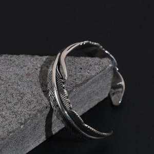 May include: A silver feather cuff bracelet. The bracelet is open and has a simple design.
