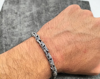 May include: A silver bracelet with an ornate, linked design. The bracelet features a series of interconnected, detailed links with dark accents. The bracelet is worn on a wrist, showcasing its intricate pattern and craftsmanship.