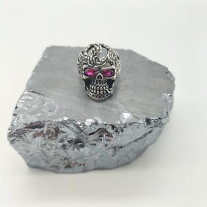 Op de afbeelding: Een zilveren schedelring met ingewikkelde details en roze edelsteenogen.