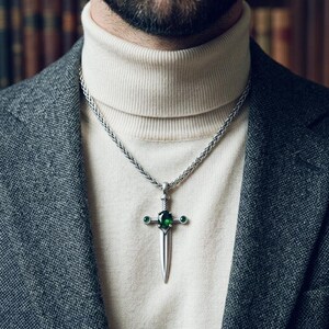 Può includere: Collana con pendente in argento con design a spada e pietre preziose verdi. La collana è indossata sopra un dolcevita color crema e un blazer grigio. Il pendente a spada ha una gemma verde centrale.