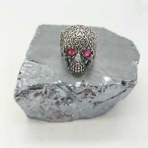 May include: A silver skull ring with intricate filigree detailing and two red gemstone eyes.