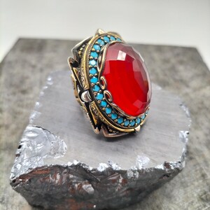 May include: A detailed ring featuring a large, faceted red gemstone. The ring has a gold-colored band with turquoise accents and intricate detailing. The ring is set on a gray, textured surface.