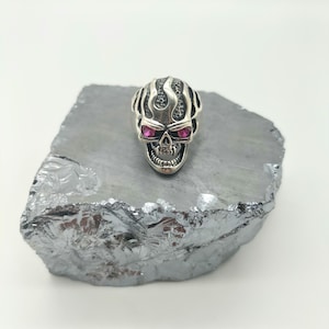 Puede incluir: Anillo de calavera de plata con ojos de gemas rosas y un diseño de llama. El anillo está sobre una roca gris.
