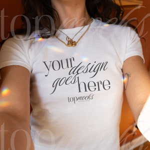 May include: White t-shirt with the text "your design goes here" in a script font. The shirt is paired with blue denim jeans. A gold necklace and hoop earrings are visible. The shirt is from topmocks.