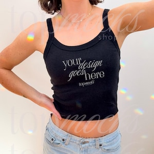 May include: A black cropped tank top with the text "your design goes here" in white. The tank top is being worn by a person with light skin and dark hair. The person is wearing blue jeans.