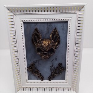 May include: A framed artwork featuring a gold-colored bat skull sculpture on a gray background. The frame is white with gold accents.