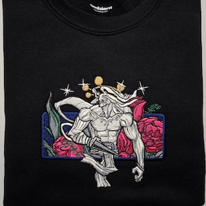 May include: A black crewneck sweatshirt featuring an embroidered graphic of a muscular, pale humanoid figure with horns, holding a sharp blade. The design includes pink roses, green leaves, and gold star accents within a blue rectangular border.