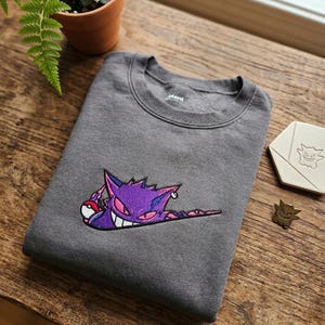 Könnte beinhalten: Ein anthrazitfarbenes Sweatshirt mit Rundhalsausschnitt und einer violetten Gengar-Stickerei. Das Stickmotiv zeigt den Charakter in einer geschwungenen Form, der einen rot-weißen Pokéball hält. Das Sweatshirt hat einen gerippten Kragen und Bündchen.